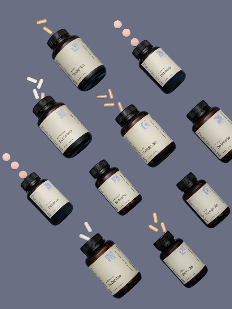 Nine amber supplement bottles with cream labels and black caps are arranged diagonally on a slate blue background, with small pink and beige capsules floating near them.